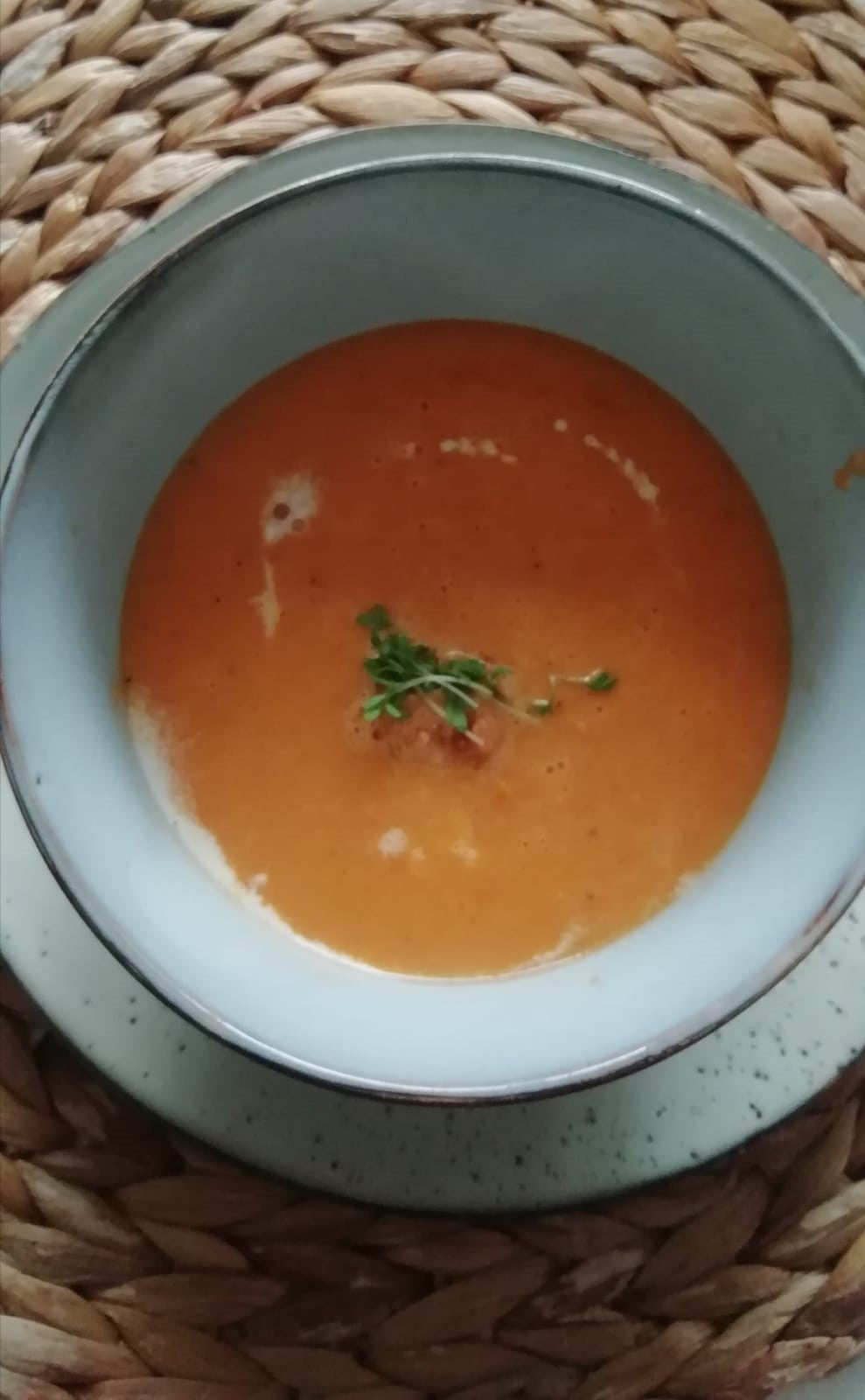 Een blauw grijze soepkom op een bijpassend bord. Hierin zit oranje tomatensoep met een room cirkel er om heen. In het midden een plukje groen.