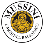 Een rond wit logo van Mussini. In het midden een geel bruine tekening van een beschermheilige.  
