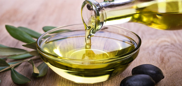 Olijfolie dat uit een glazen fles in een schaap wordt gegoten. Schaal staat op een houten tafel. Op de tafel liggen olijven en een olijftak. 